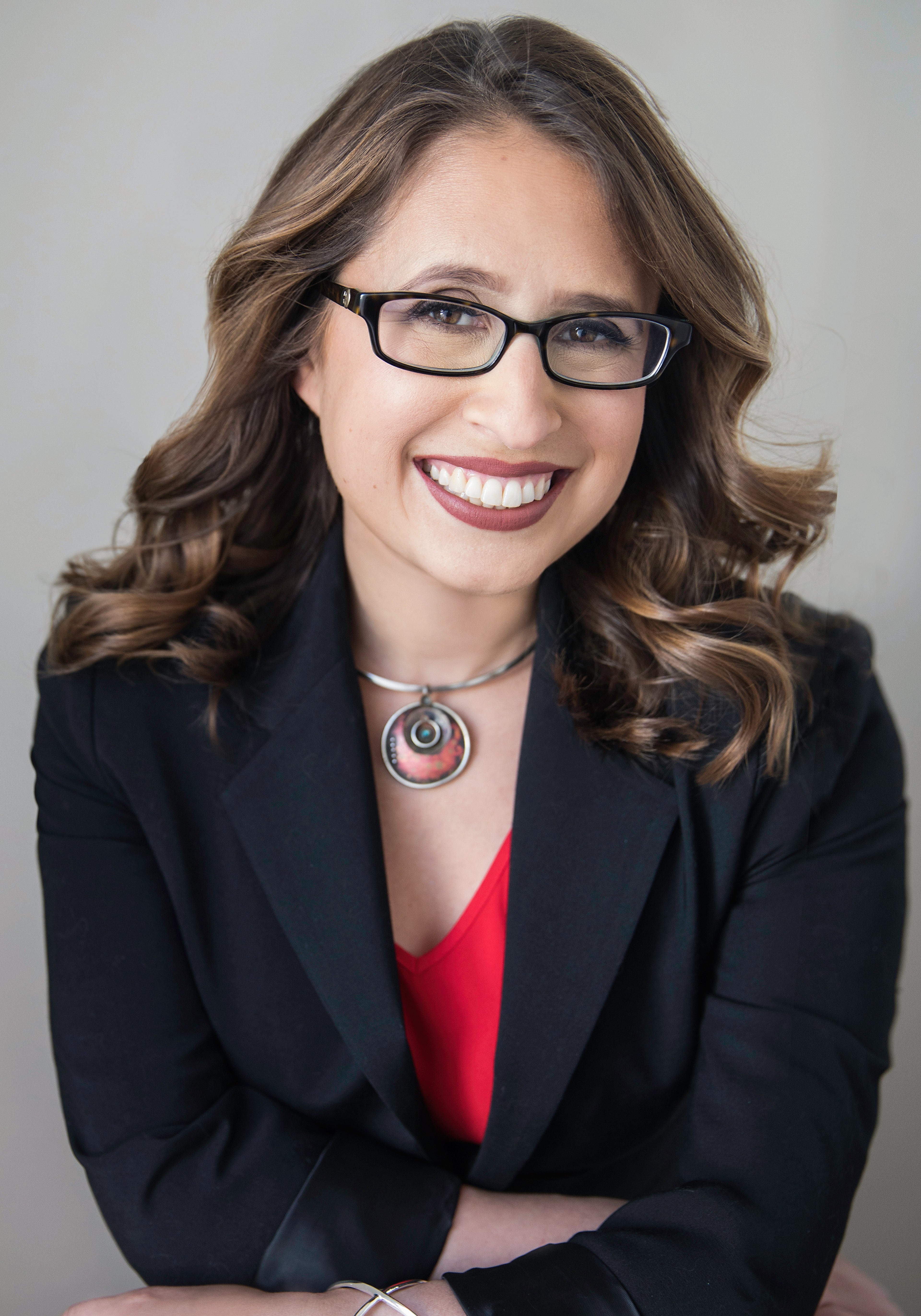 Woman wearing glasses and a black blazer with a red top against a gray background
