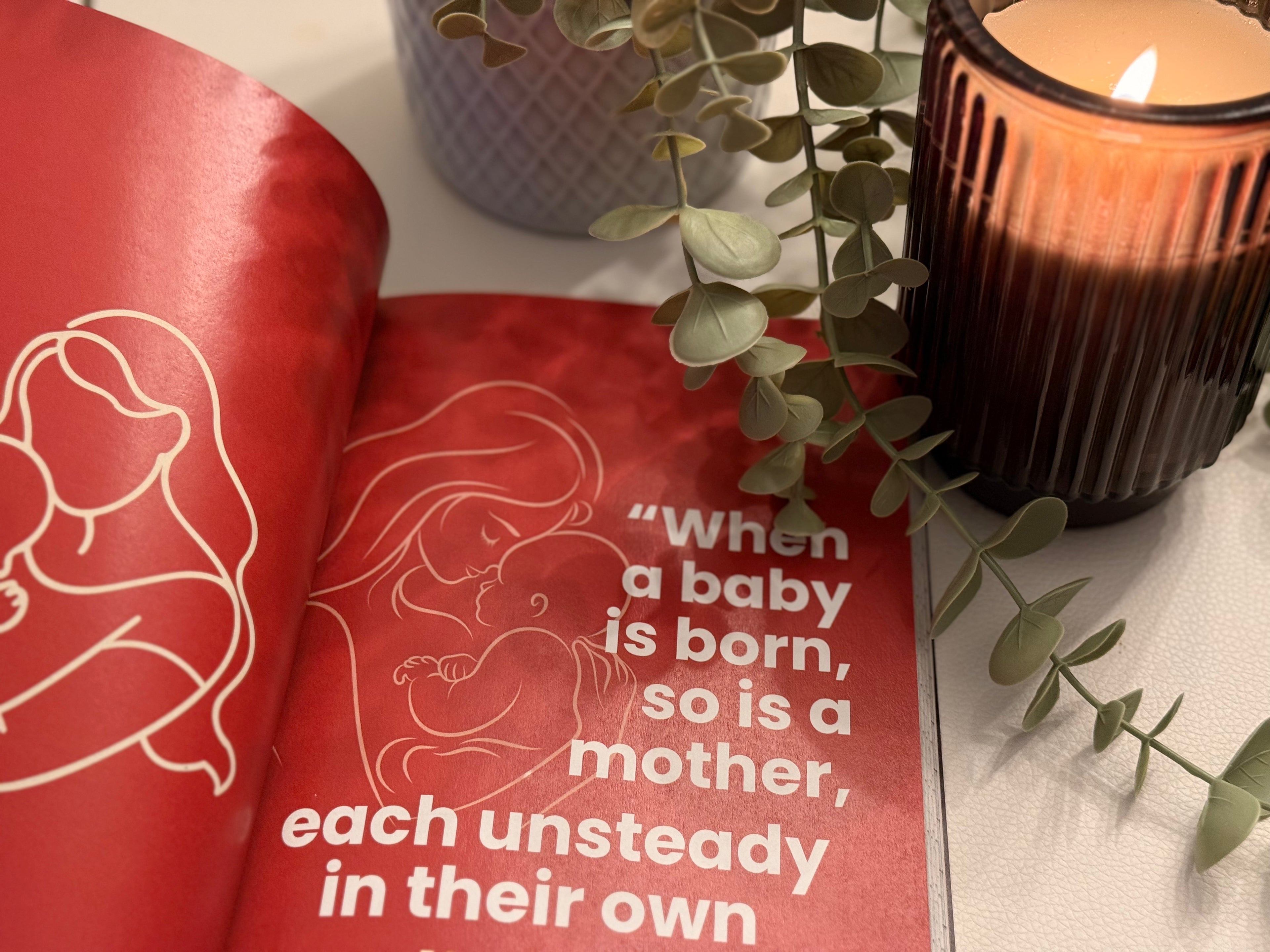 Open book with a quote about a mother and baby, next to a lit candle and plant on a white surface.